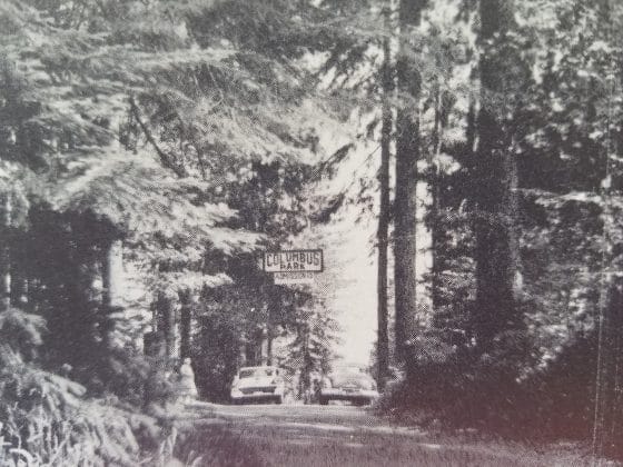 A black-and-white photograph shows cars on a wooded lane beneath a Columbus Park sign.