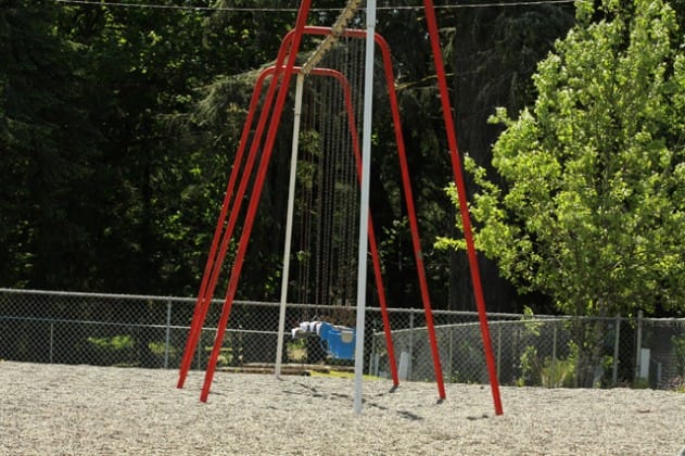A red swing frame stands in a gravel play area bordered by trees.